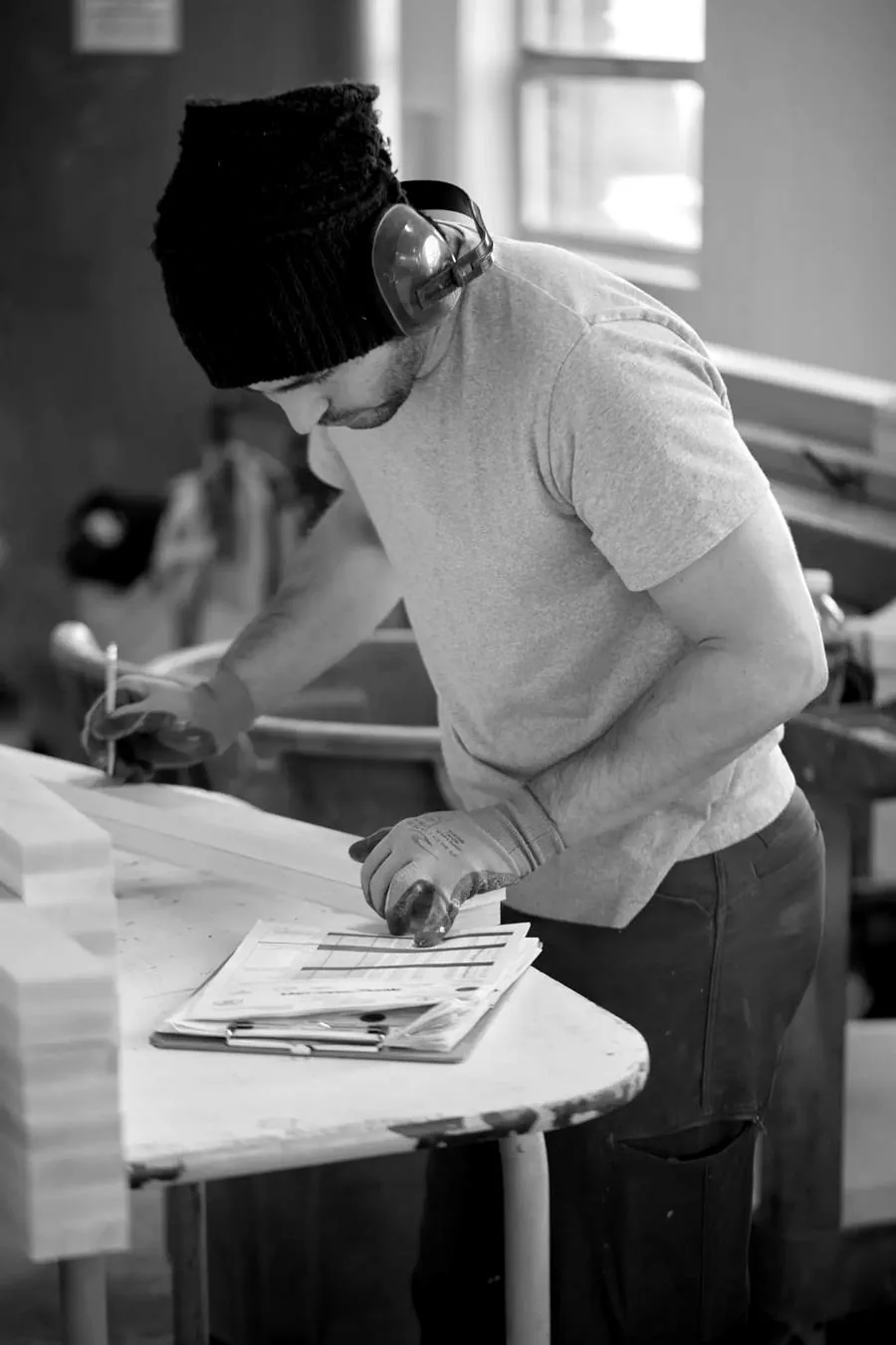 A craftsman in a knit cap and ear protection marks a stack of door parts at the bench